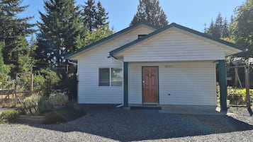 A small, single-story building with a brown door and a window, surrounded by gravel and trees.