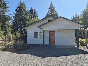 A small, single-story building with a brown door and a window, surrounded by gravel and trees.