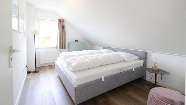 A bedroom with a bed, a bedside table, a lamp, a radiator, and a window with curtains. - Stylish Waterfront Villa With Sauna (Makkum)