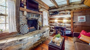 Interior de una cabaña de troncos con chimenea de piedra, un televisor montado encima y una zona de estar acogedora con un sofá de cuero y una mesita.