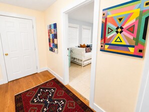 Un couloir avec une porte blanche, une peinture abstraite aux couleurs vives, un tapis à motifs et, au fond, un aperçu d’une chambre à coucher avec un lit blanc et une fenêtre équipée de stores.