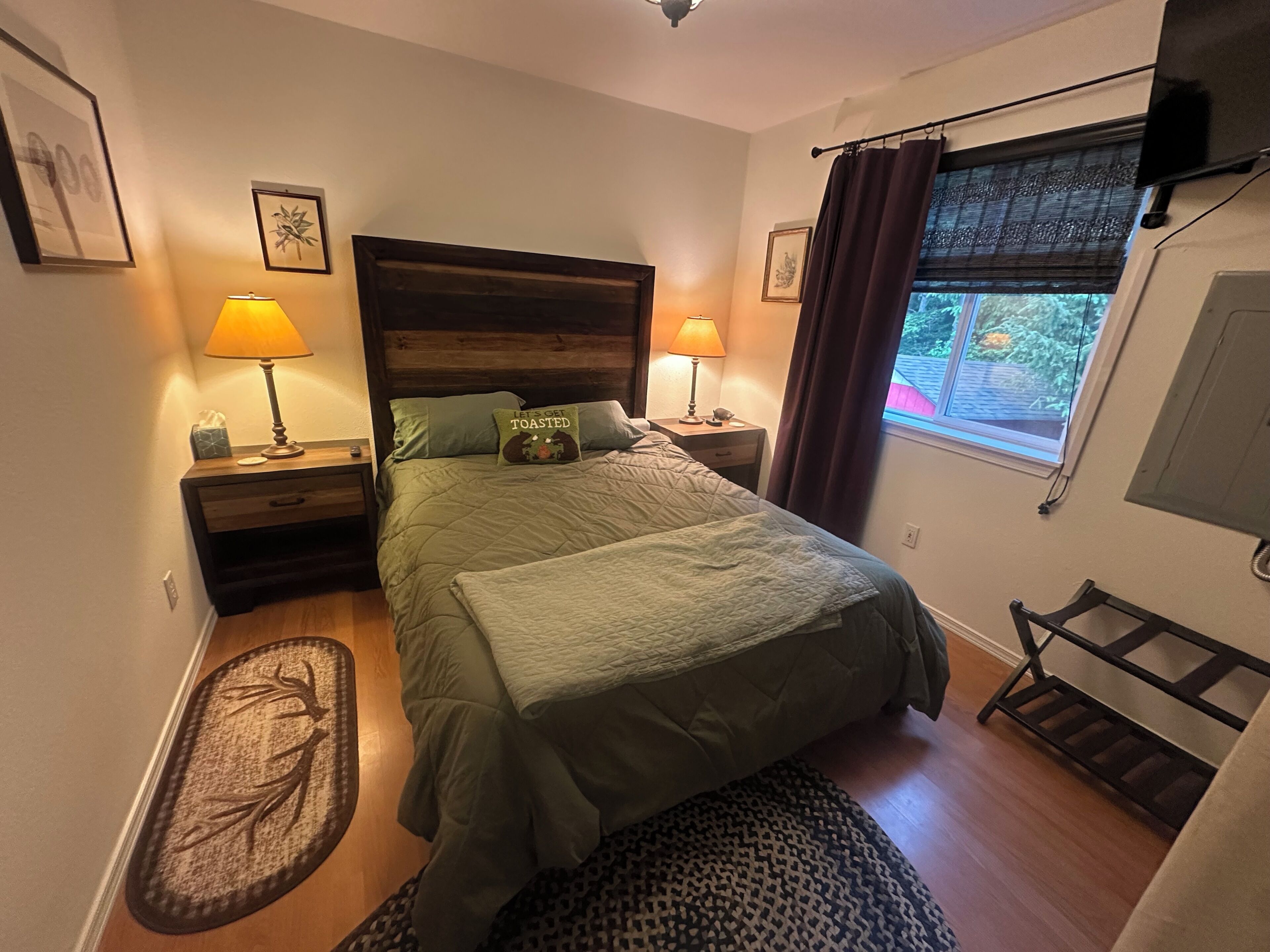 Une chambre à coucher comprenant un lit, deux lampes de chevet, une fenêtre avec des rideaux, une table de chevet avec une lampe et un téléviseur fixé au mur.