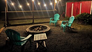 An outdoor seating area with chairs and a fire pit, surrounded by a red building and string lights.