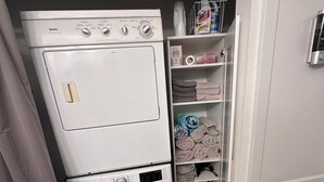 A compact laundry area with a front-loading washing machine and dryer, a shelf with folded towels, and a tiled floor.