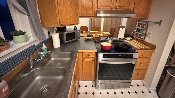A compact kitchen with wooden cabinets, a stainless steel sink, a microwave, an oven, and a black and white tiled floor.