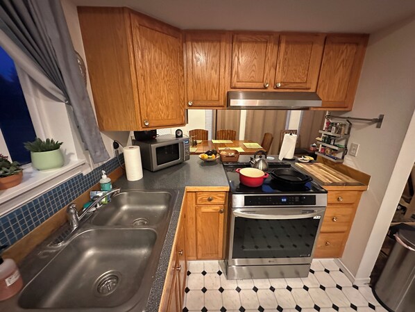 A compact kitchen with wooden cabinets, a stainless steel sink, a microwave, an oven, and a black and white tiled floor.