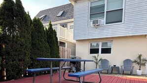 Eine rote Holzterrasse mit einem Metall-Picknicktisch und -stühlen, umgeben von einem Wohngebäude mit Satteldach und eingezäuntem Garten.