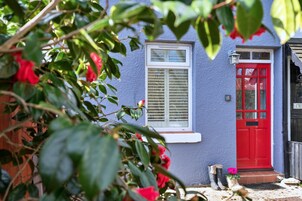 Une maison avec une porte rouge, une fenêtre avec des volets et un petit jardin agrémenté de plantes et d’une paire de bottes.
