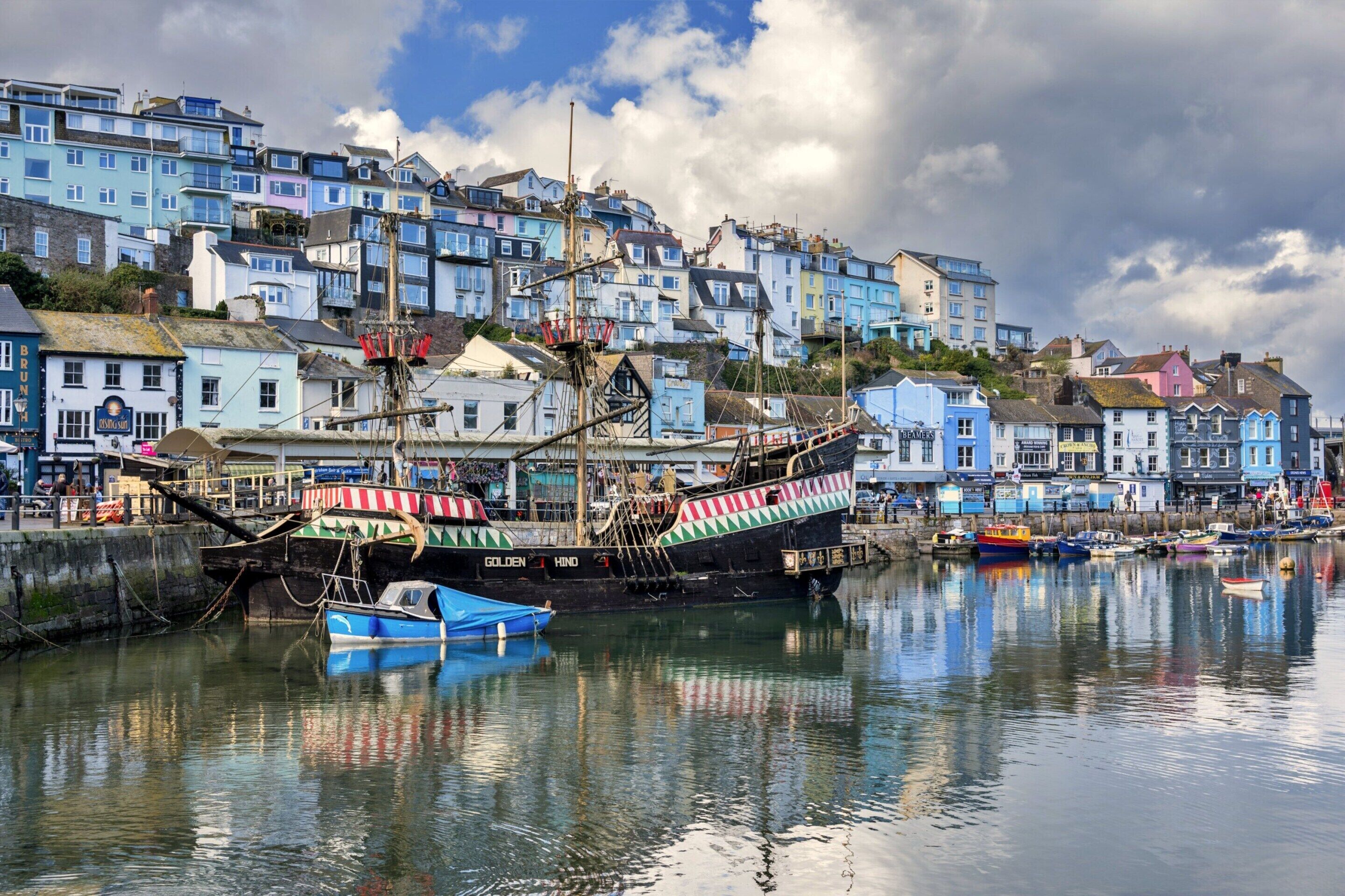A harbor with a large sailing ship docked, surrounded by colorful buildings and calm water reflecting the scene.