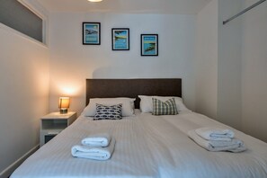 Ein ordentlich bezogenes Bett mit weißen Leinen und zwei Kissen, eine Nachttischlampe sowie drei gerahmte Bilder an der Wand.