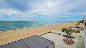 Une vue sur la plage depuis un balcon, avec une table et des chaises, un petit jardin et un ciel dégagé.