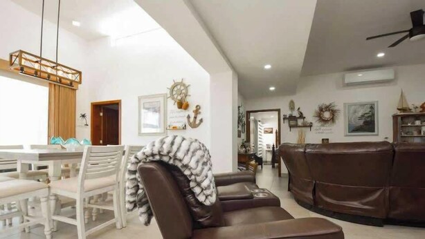 Un salon moderne avec un canapé en cuir marron, un coin repas avec des chaises blanches et un ventilateur de plafond.