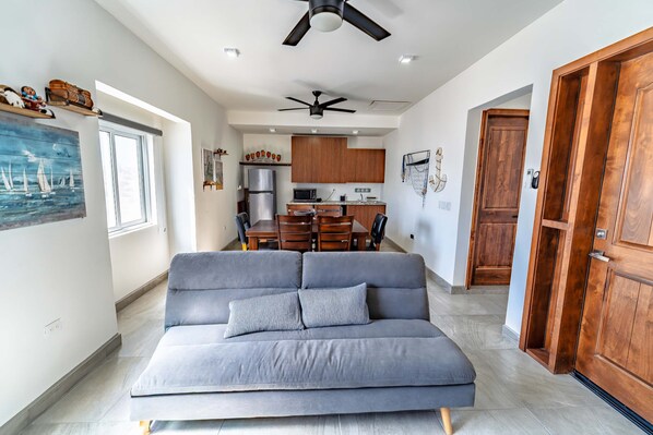 Ein modernes Wohnzimmer mit einem grauen Sofa, einem Deckenventilator und einer Küche im Hintergrund.
