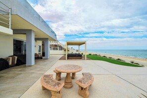 Eine Terrasse mit rundem Tisch und Bänken, einer überdachten Sitzgelegenheit und Blick auf den Strand.