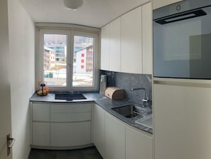 Ett kompakt kök med en inbyggd ugn, en diskho och ett fönster med utsikt över byggnaderna utanför.