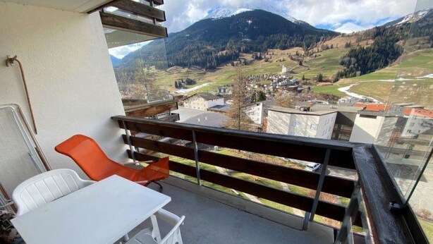 Ein Balkon mit Tisch und Stühlen, mit Blick auf eine gebirgige Landschaft mit Gebäuden.