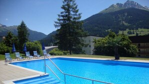 Ein Swimmingpool mit klarem, blauem Wasser, umgeben von Liegestühlen und Sonnenschirmen, mit Blick auf eine Berglandschaft.