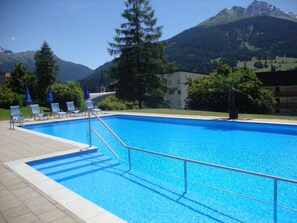 En pool med kristallklart blått vatten, omgiven av solstolar och parasoller, med ett berg i bakgrunden.