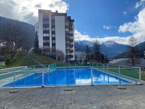 A swimming pool with a view of a mountain range and a modern building in the background. - Tgesa Piz Mez 24 Appartement/Fewo, Dusche, WC, 2 Schlafräume by Interhome (Savognin)
