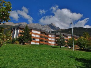 A multi-story hotel with a flat roof, surrounded by a well-maintained lawn and trees, with mountains in the background. - Tgesa Sunnmatt Nr 46 Appartement/Fewo, Dusche, WC, 1 Schlafraum by Interhome (Savognin)