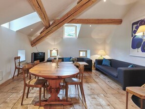 Ein geräumiger Loft mit hoher Decke, freiliegenden Holzbalken, einem großen runden Holztisch und einer gemütlichen Sitzecke mit Sofa und Sesseln.