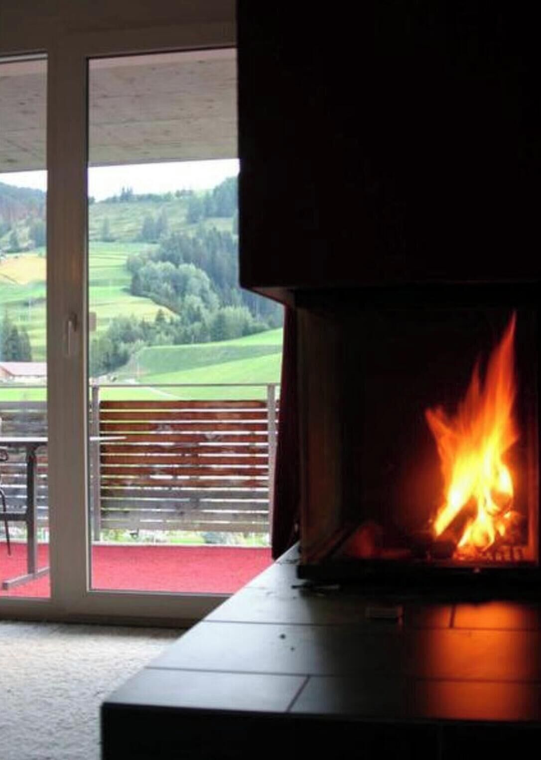Ein Kamin mit sichtbarem Feuer, eine Schiebetür zu einem Außenbereich mit Blick auf eine grüne Landschaft.
