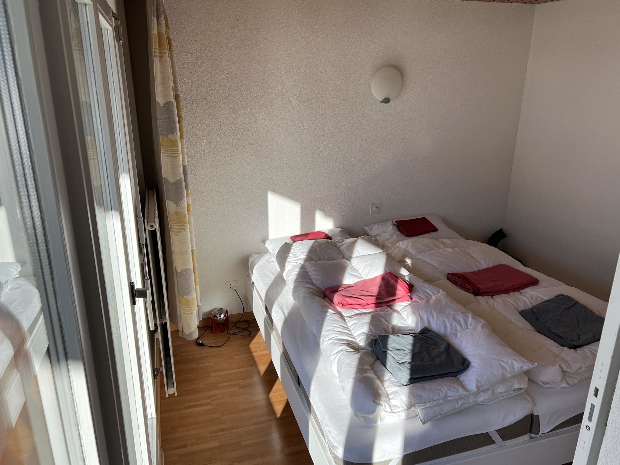 En säng för en person med vita sängkläder och röda kuddar, ett eluttag och en väggmonterad lampa.