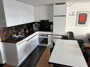 A compact kitchen with white cabinets, a black countertop, and a dining area with a white table and black chairs. - Som Barnagn 7 Appartement/Fewo, Bad, WC, 3 Schlafräume by Interhome (Savognin)