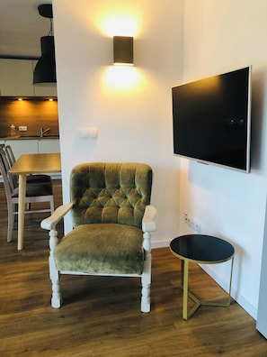 A living room with a tufted armchair, a flat-screen TV mounted on the wall, a small round table, and a wooden dining table with chairs. - Panoramic Apartment in the Black Forest, Prime Location, Terrace, Garden, Near Baden-Baden (Bühlertal)