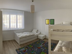 A child's bedroom with a bunk bed, a single bed, a window with blinds, a bedside table, and two framed pictures on the wall.