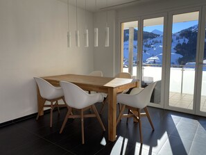 Ein moderner Essbereich mit einem Holztisch und weißen Stühlen, große Fenster mit Blick auf die Berge.