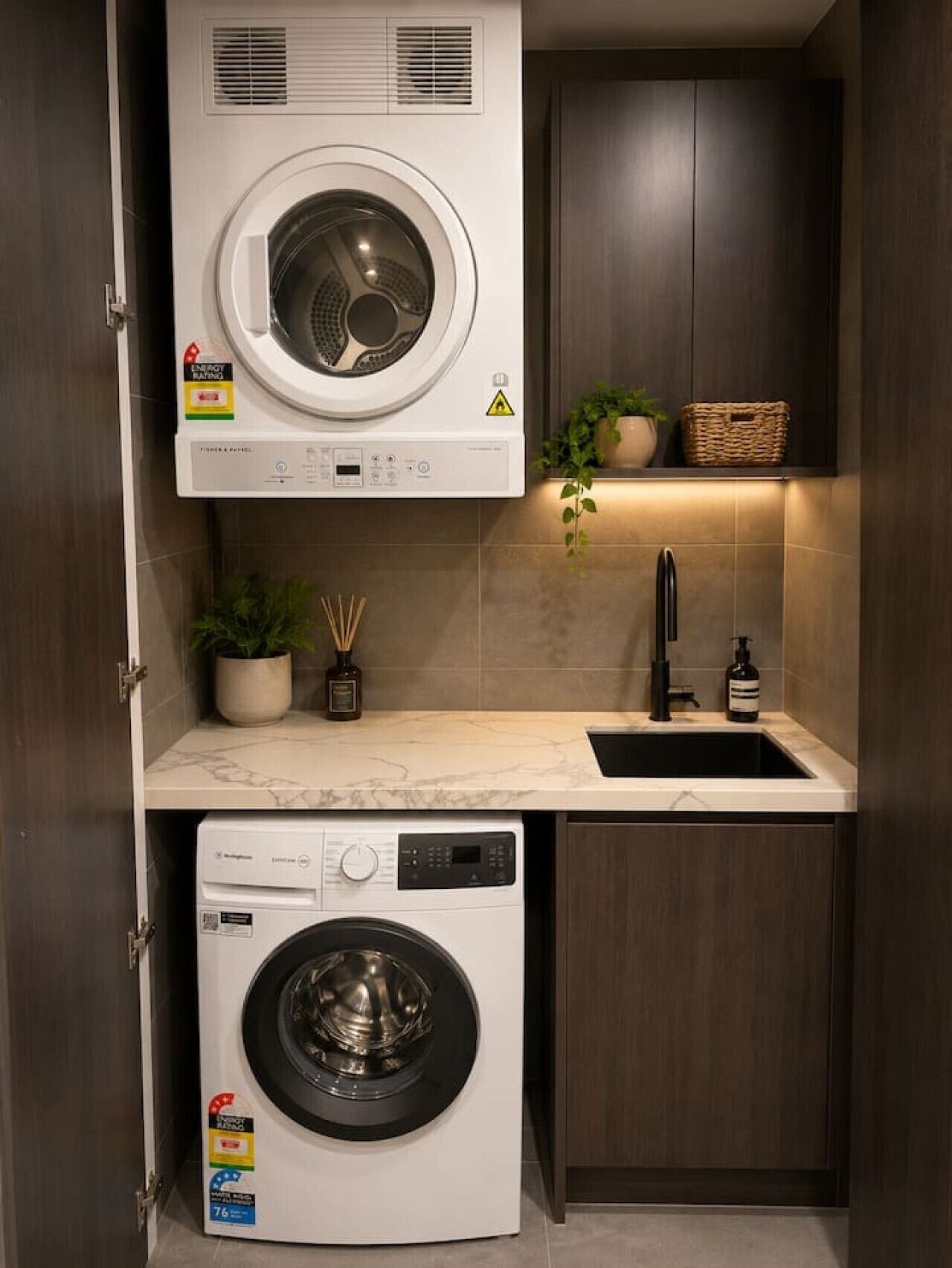 A compact laundry area with a front-loading washing machine and a dryer, a sink with a faucet, and a small shelf with a plant and a basket.
