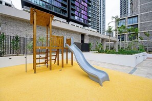 A playground with a slide and climbing structures, surrounded by a white wall and tall buildings. - Spacious Pool Retreat with Gym Pool BBQ Rooftop (Lidcombe)