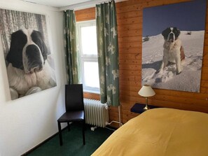 A hotel room with a bed, a chair, a radiator, a window with curtains, and two large dog portraits on the wall. - Haus Viols 54 Appartement/Fewo, Bad, WC, 2 Schlafräume by Interhome (Savognin)