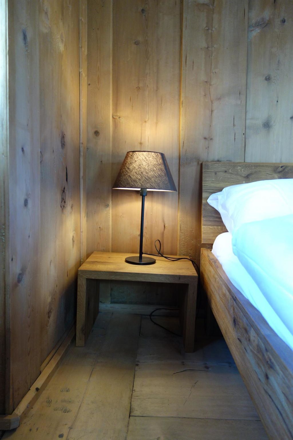 A wooden bedside table with a lamp, a bed with white bedding, and wooden walls.