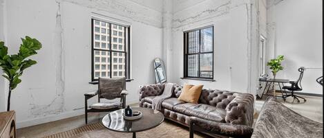 A room with a leather sofa, a round coffee table, and a chair. There are two windows with metal grilles, a plant, and a mirror on the wall.