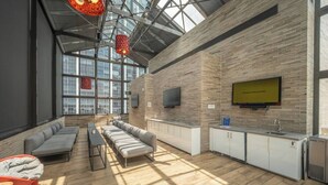 A modern interior with a high ceiling, a seating area with sofas and a table, a kitchen area with a sink and cabinets, and a large window providing natural light. - Penthouse Loft, Central Downtown Dallas, Pool+Gym+Office, City Views (Dallas)