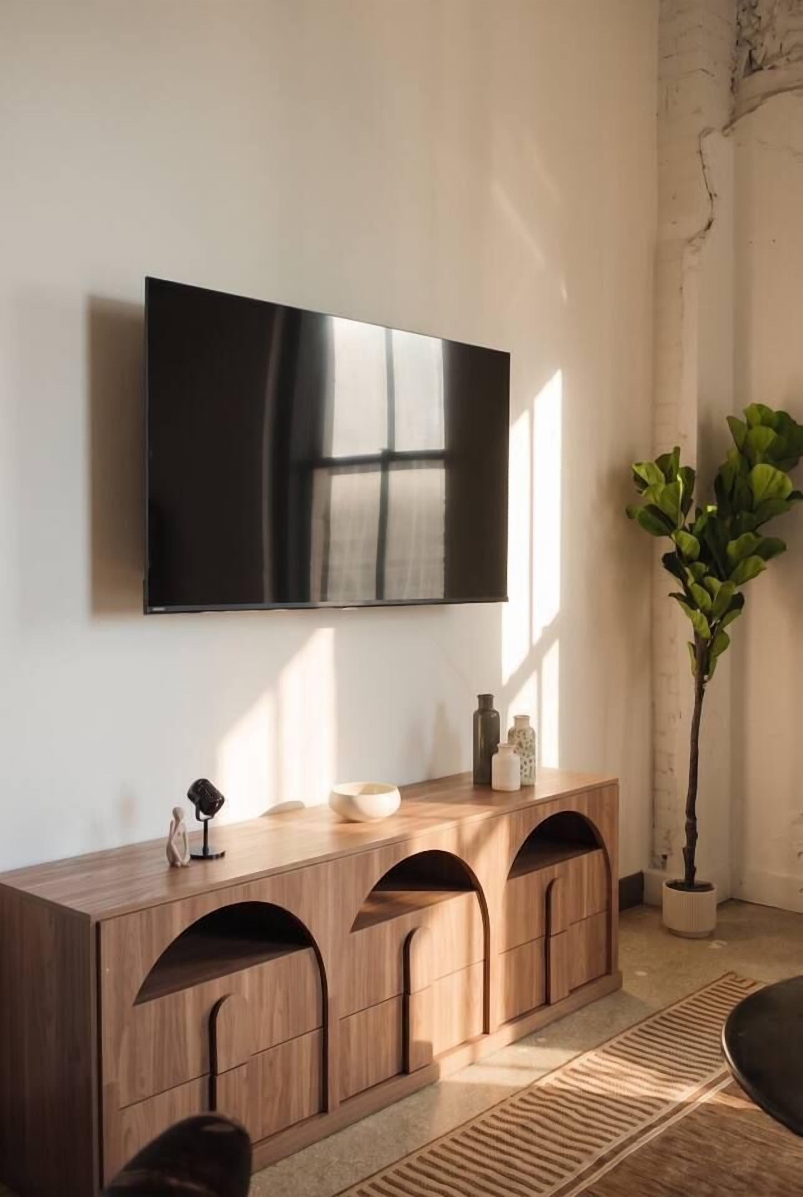 A modern living room with a flat-screen TV mounted on the wall, a wooden console table with drawers, a potted plant, and a patterned rug.