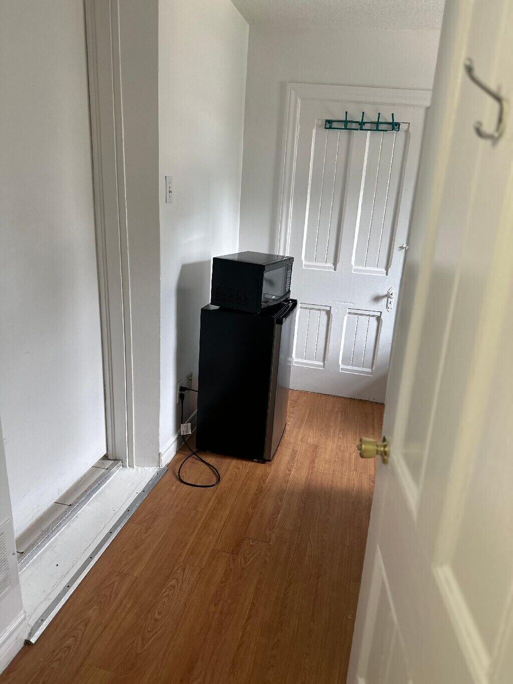 Un couloir étroit avec un réfrigérateur noir et un four à micro-ondes placés l’un sur l’autre, une porte blanche avec un porte-manteau, et un plancher en bois.