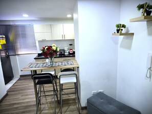 Eine kleine Küche mit Esstisch, Stühlen, einer Blumenvase, einem Kühlschrank und einer Kaffeemaschine.