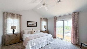 A bedroom with a large bed, two bedside tables with lamps, a ceiling fan, and a view through a sliding glass door. - 1822 - Our Happy Place By The Sea, Corolla Retreat | Saltwater Pool & Arcade Game (Corolla)