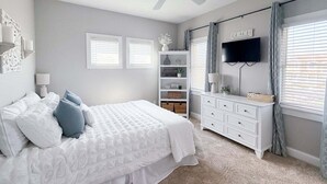 A bedroom with a bed, a dresser, a TV, and a shelf with decorative items. - 1822 - Our Happy Place By The Sea, Corolla Retreat | Saltwater Pool & Arcade Game (Corolla)