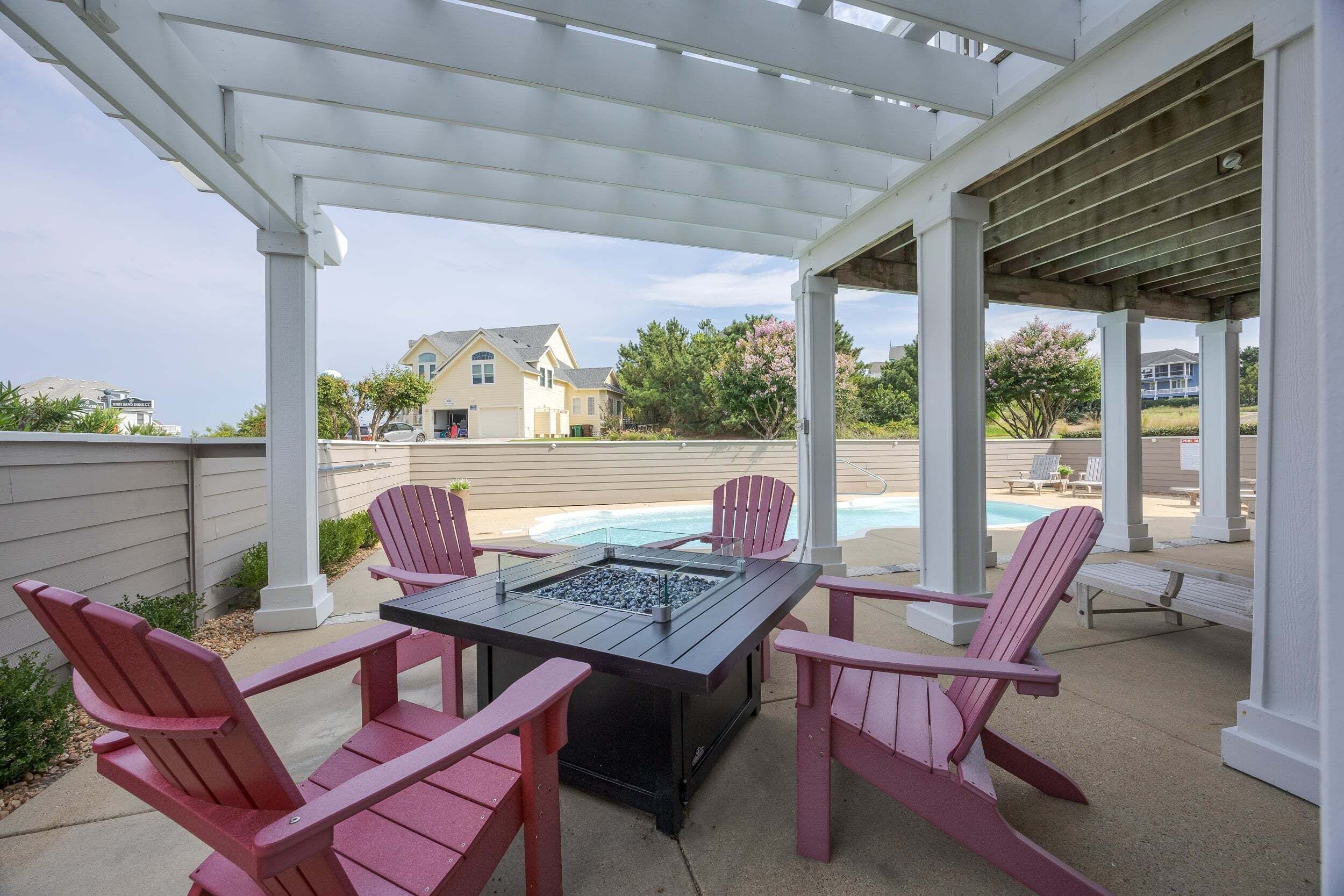 A covered outdoor seating area with Adirondack chairs and a fire pit, overlooking a swimming pool and a residential neighborhood.