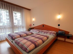 A double bed with a plaid bedspread, two bedside tables with lamps, a window with sheer curtains, and a view of buildings outside. - GOLF Apartments Appartement/Fewo 6-Bettwohnung Nr. 1 by Interhome (Saas-Fee)