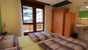 A hotel room with two beds, a window with a view of buildings, a wooden wardrobe, and a bathroom with a sink. - GOLF Apartments Appartement/Fewo 4-Bettwohnung Nr. 3 by Interhome (Saas-Fee)