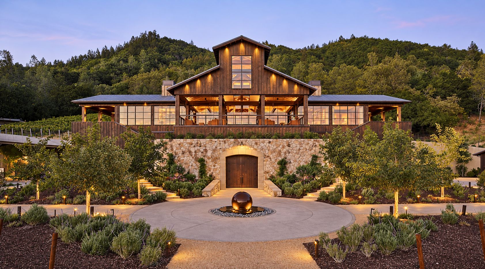 A large, two-story wooden building with a stone facade, a central entrance, and a landscaped driveway with a spherical sculpture.