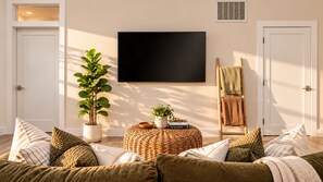 A living room with a flat-screen TV mounted on the wall, a potted plant, a woven ottoman, and a wooden chair with a blanket. - Casa Solara: Modern Luxury. Spa. Fire Pit - Minutes to Downtown Napa (Napa)