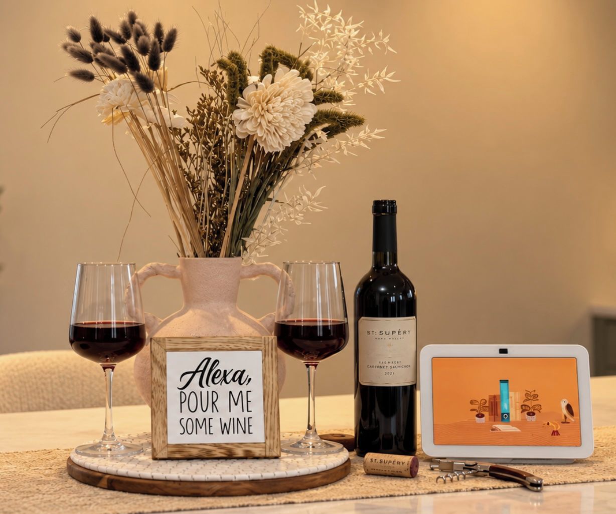 Uma garrafa de vinho, dois copos de vinho, um vaso com flores, uma placa e um tablet sobre a mesa.