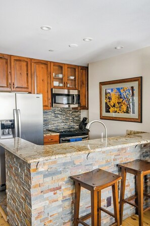 A kitchen with wooden cabinets, stainless steel appliances, a tiled backsplash, and a bar area with stools. - Upscale Condo in Downtown with Ski In Access, Pool, Hot Tubs, Fitness Center (Breckenridge)