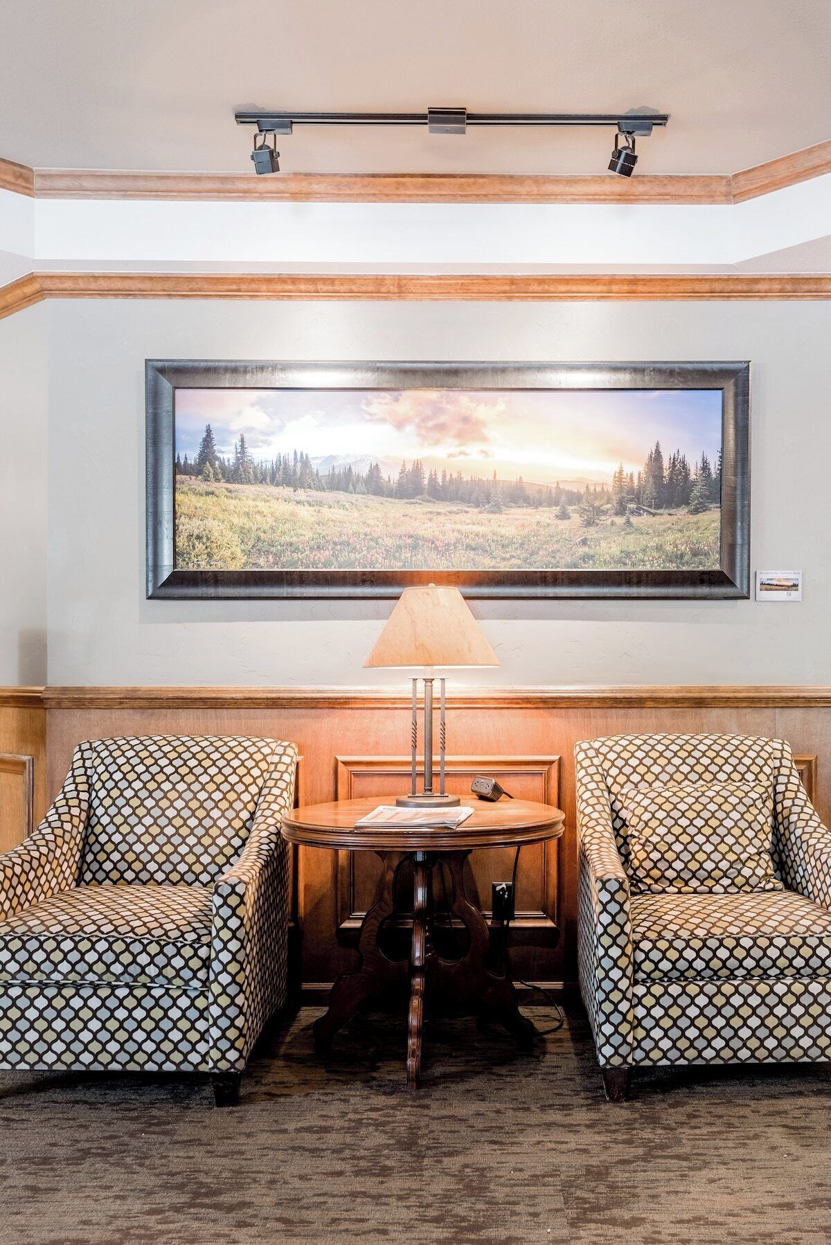 A room with a large framed landscape painting, a table with two patterned chairs, and a floor lamp.